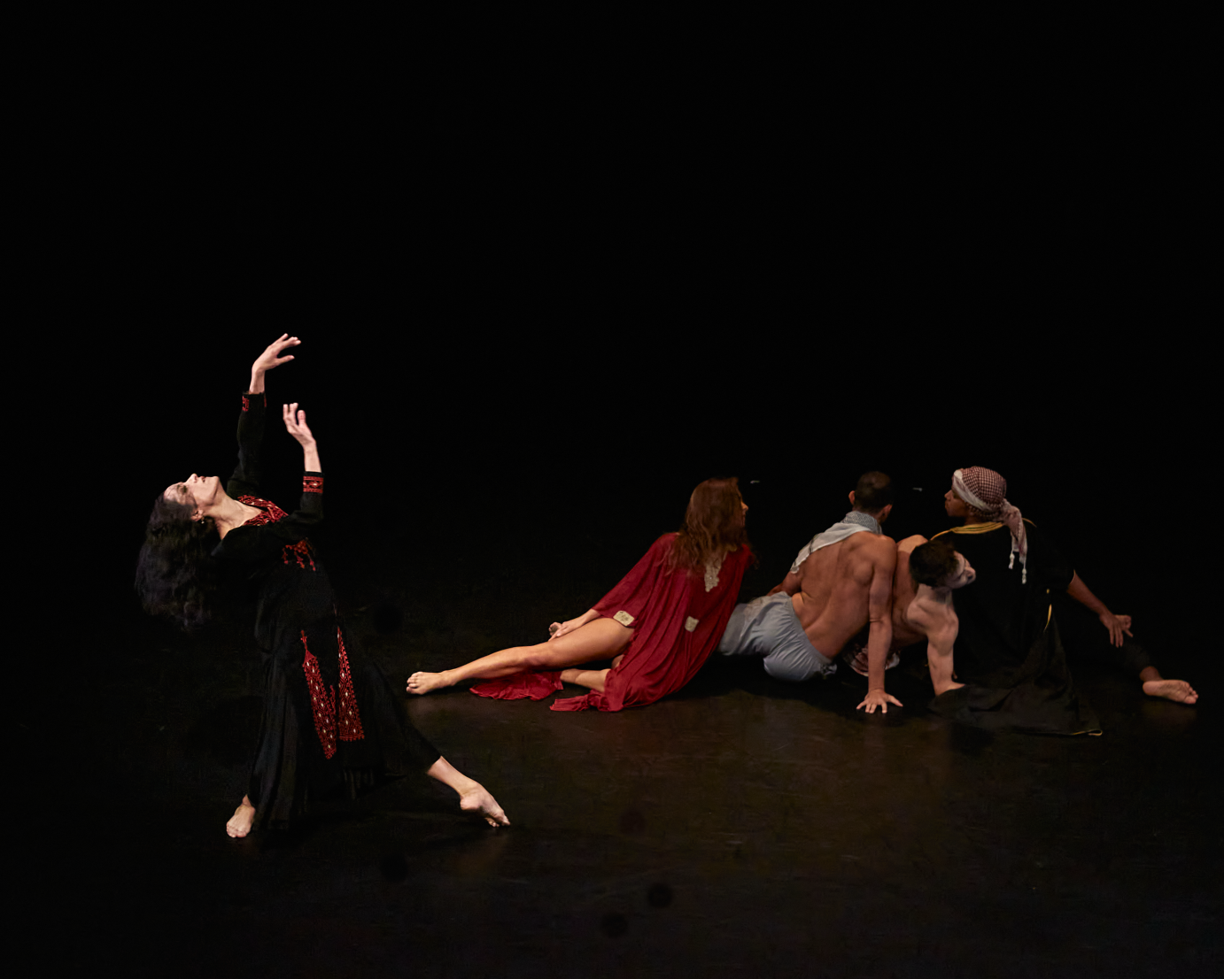 The cast of the Mirage sits on the floor in a diagonal, their backs to the audience. Elisa Toro Franks stands at the end of the diagonal. She lifts a hand and gazes upward.